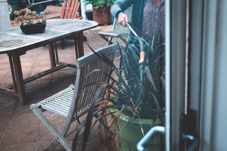 A person engaged in garden maintenance on a patio area, using a string trimmer to trim tall, dark green ornamental grass in a large planter. In the background, there is a weathered wooden outdoor dini